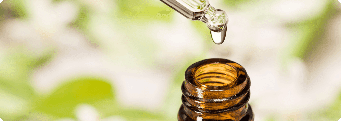 Gota de óleo essencial sendo aplicada com conta-gotas em frasco âmbar, com fundo desfocado de folhas e flores, representando o uso de aromaterapia no ambiente doméstico.