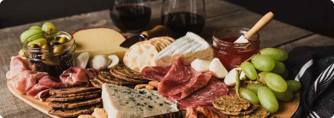 Tábua de frios completa com vinhos ao fundo, queijos variados, embutidos e frutas frescas.